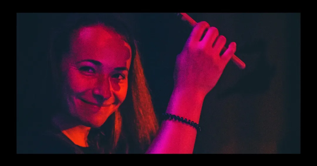 Calie, a Rock Star Brass drummer, smiling towards the camera with a drumstick raised, lit deep red on stage.