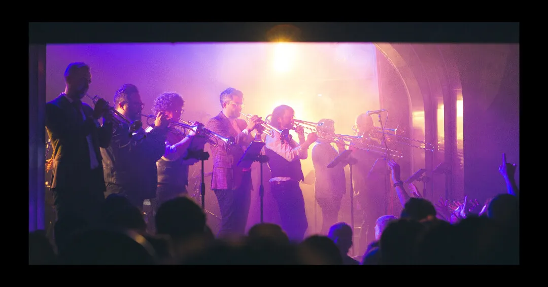 The Rock Star Brass horn line playing side by side on stage, lit by magenta haze, with the crowd's hands in the air.